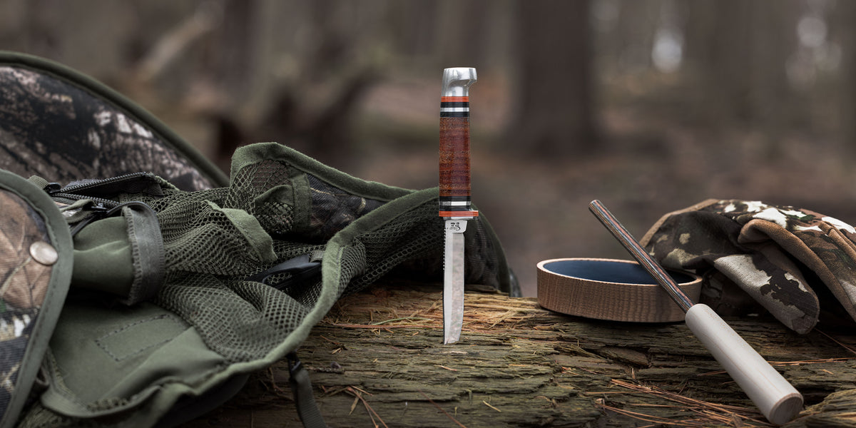 A folding knife with a brown wood-grain handle stands upright on a log in a forest setting, surrounded by camouflage gear including a backpack, gloves, and a sharpening stone.