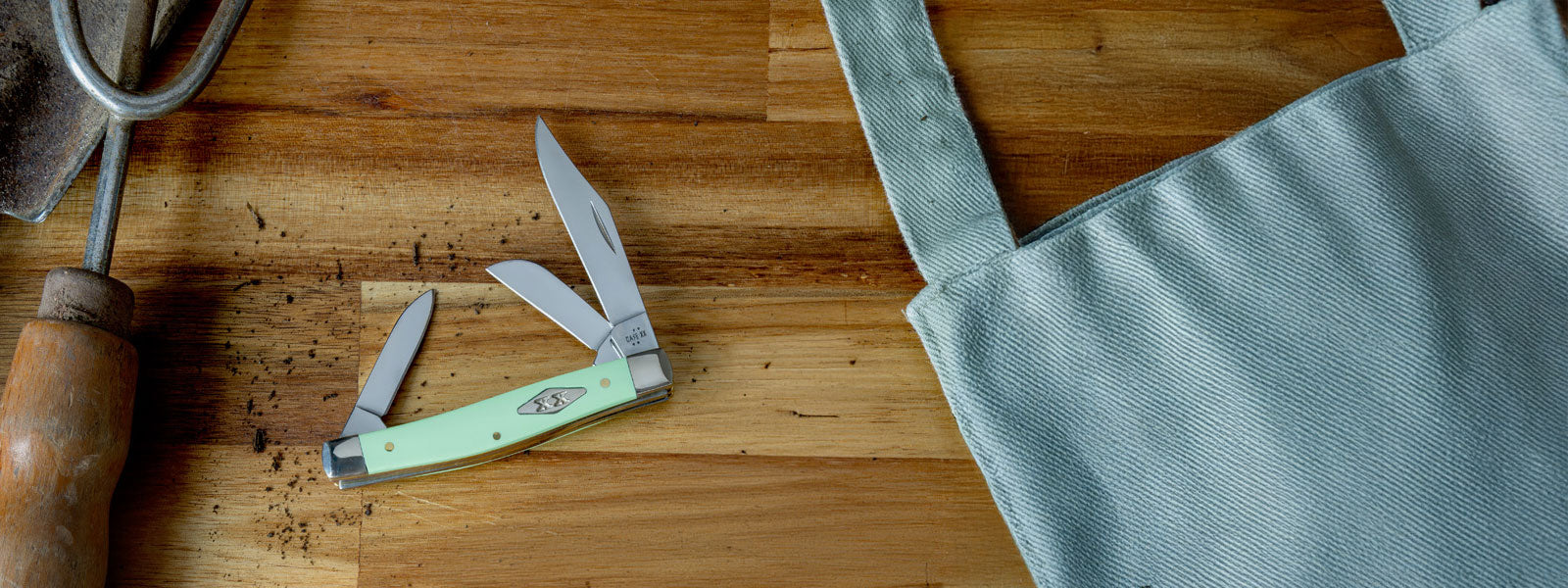 Case knife on a wooden surface with an apron and garden tool in the background.