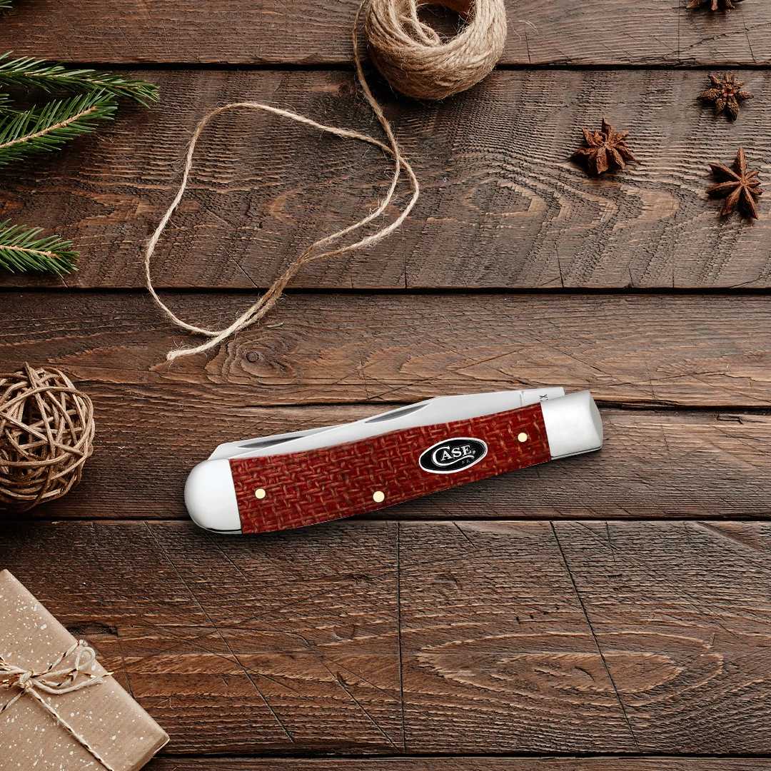 A Case knife with a red woven-texture handle lies on a dark wooden surface, surrounded by holiday-themed props including pine branches, twine, star anise, a ball of twine, and a wrapped gift box.
