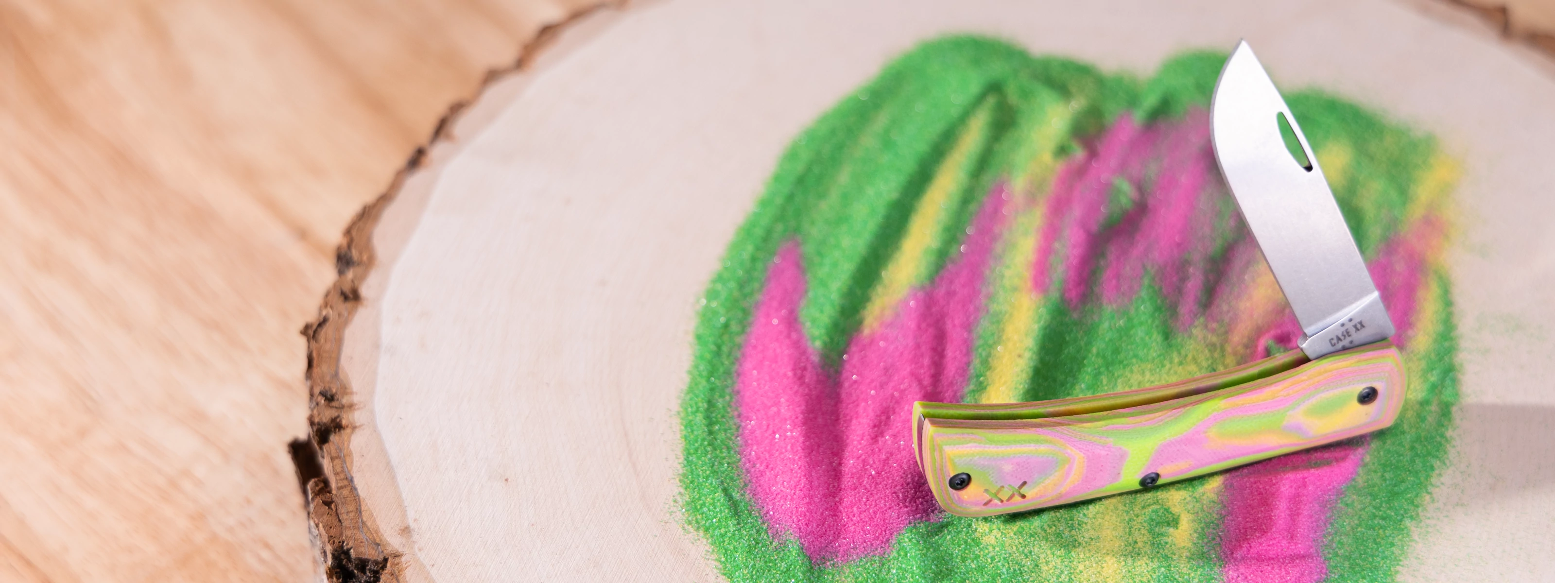 A Case knife with a metallic pink and green handle and a silver blade, resting on a colorful green and pink painted wood slice over a natural wood surface.