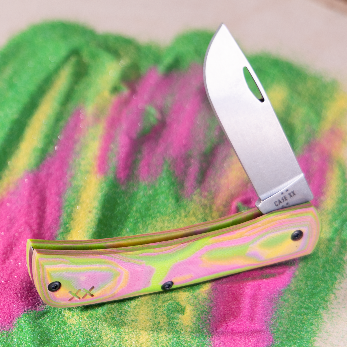 A Case knife with a metallic pink and green handle and a silver blade, resting on a colorful green and pink painted wood slice over a natural wood surface.