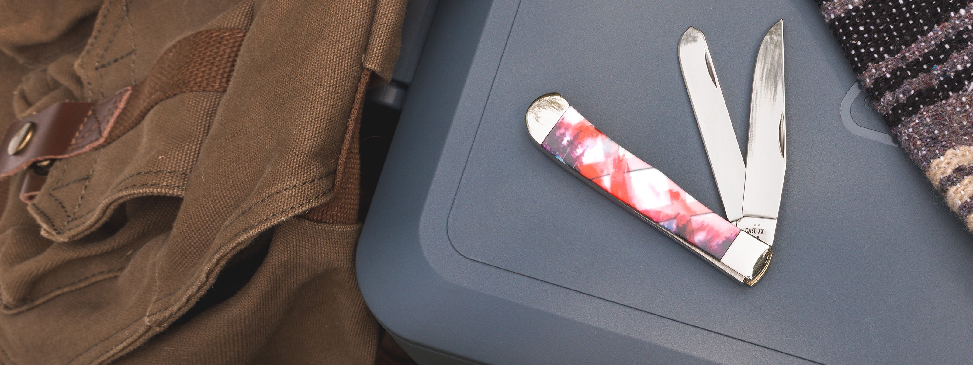 A Smooth Color Infusion XX® Mountain Trapper is shown open on a dark gray hard case, surrounded by a brown canvas bag on the left and a patterned textile on the right.