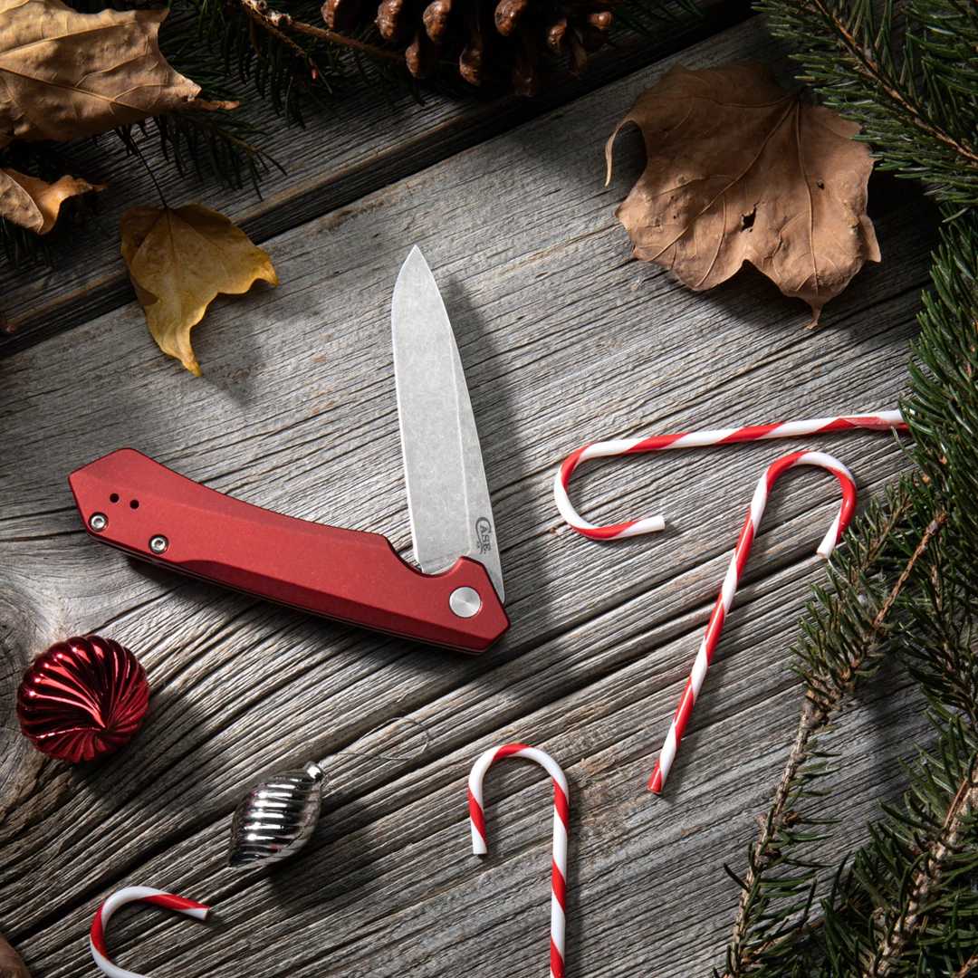 A red Case knife with a silver blade is displayed on a weathered gray wooden surface, surrounded by Christmas-themed props including candy canes, pine branches, pinecones, and dried leaves.