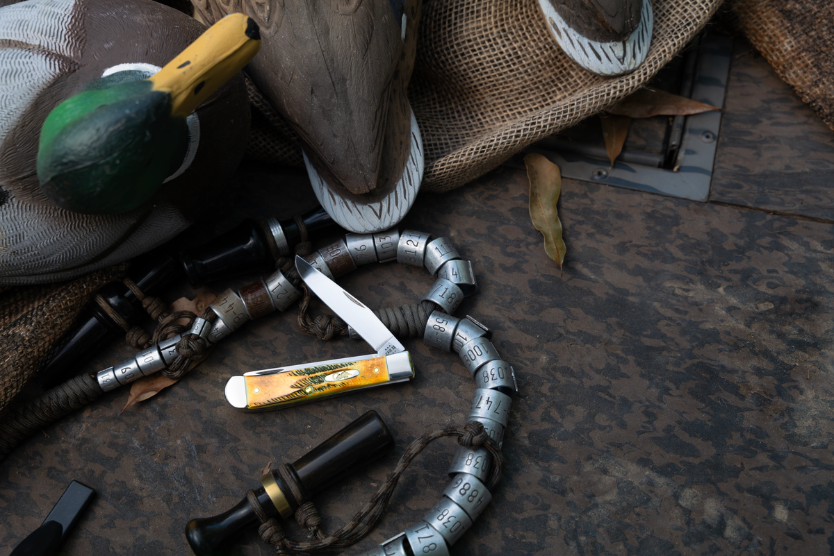 Duck hunting accessories arranged on a dark textured surface, including a Case knife and a duck call.
