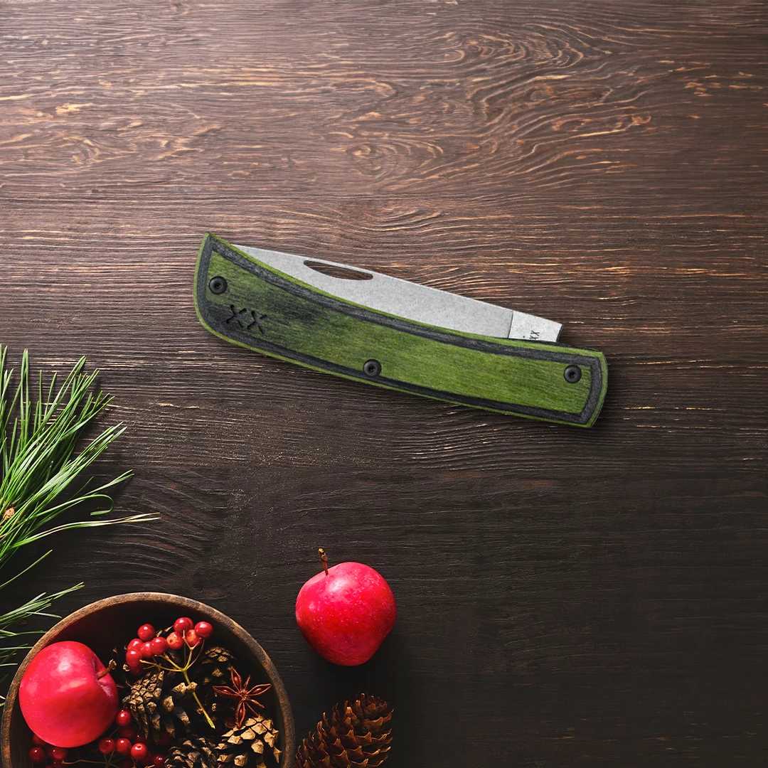 A Case pocket knife with a textured green handle and a silver stainless steel blade, displayed diagonally on a dark brown wooden tabletop. The scene is styled with a holiday theme, featuring pine needles, a small wooden bowl with red apples, and pinecones in the lower-left corner.