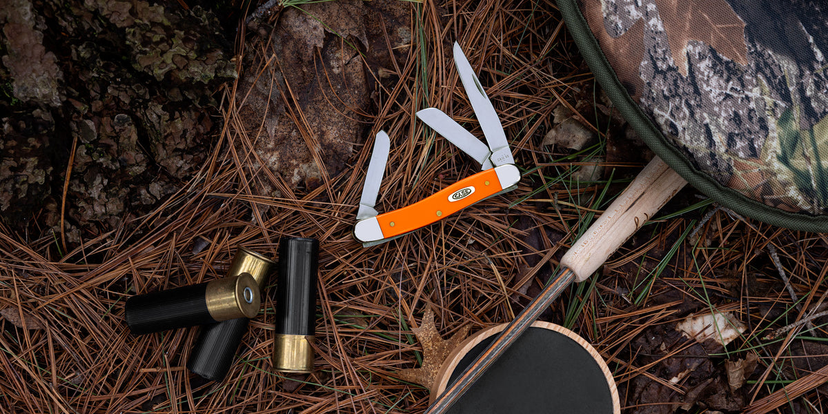 An orange folding pocket knife with white blades is displayed on a forest floor covered in pine needles, alongside shotgun shells, a bow, and other outdoor gear.