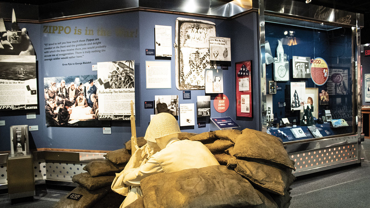 A museum display featuring a soldier mannequin in a trench with sandbags in the foreground, alongside historical exhibits on blue walls and a glass case containing memorabilia.