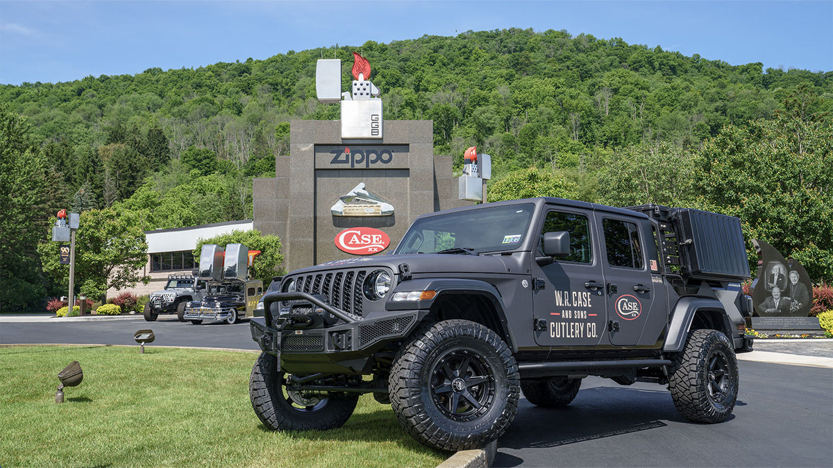 The Case Jeep is parked outdoors in front of a large Zippo/ Case Museum, with a forested hill in the background. 
