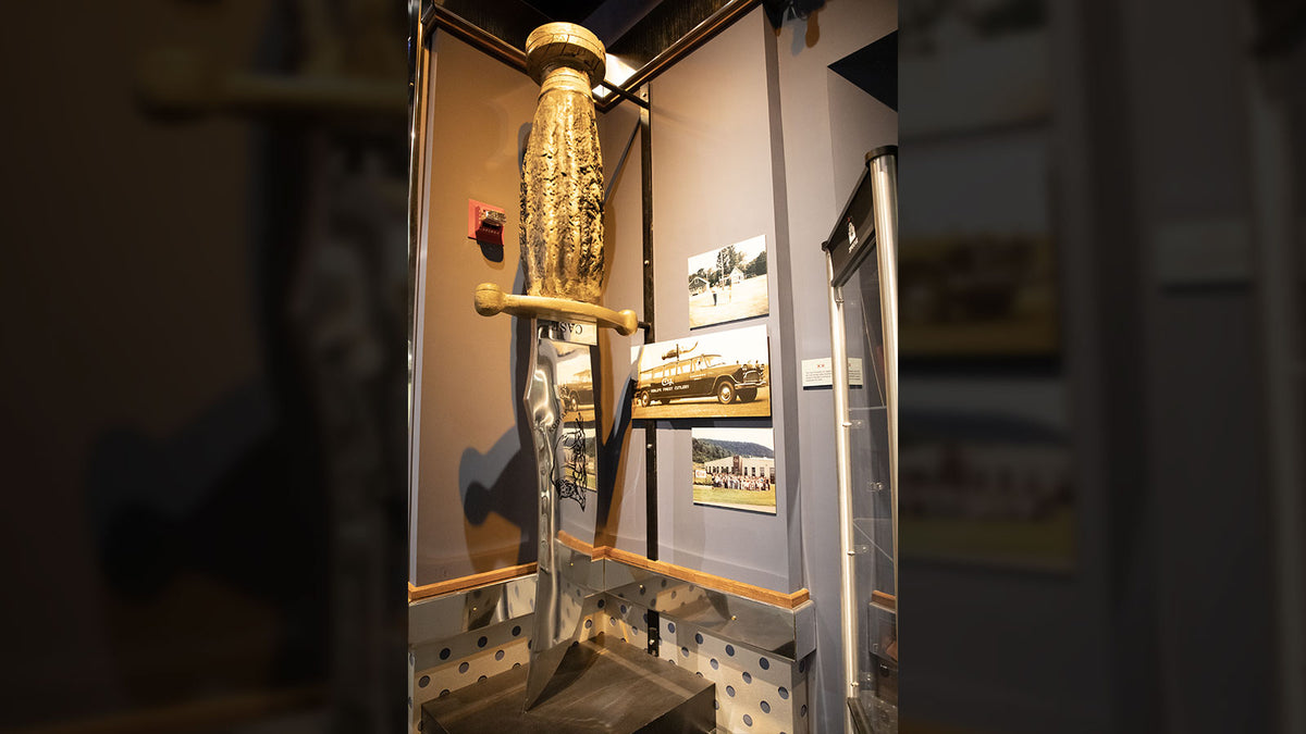 A large Kodiak knife mounted vertically inside a glass display case, shown with framed pictures on the wall.
