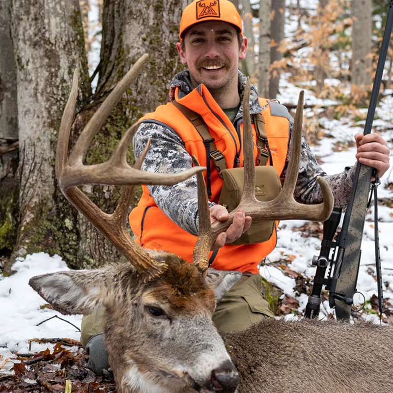 BEAU MARTONIK: In a forest with snow on the ground wearing an orange hunting hat and bright orange vest while kneeling behind a large deer with antlers and holding a rifle.