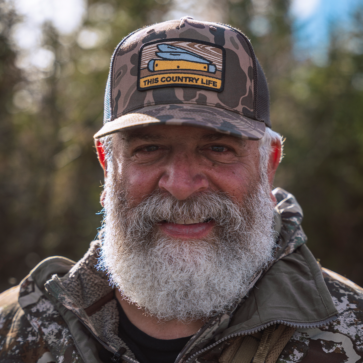BRENT REAVES: Wearing a camouflage trucker hat with a 'This Country Life' patch and a matching camouflage jacket, set against a blurred outdoor forest background.