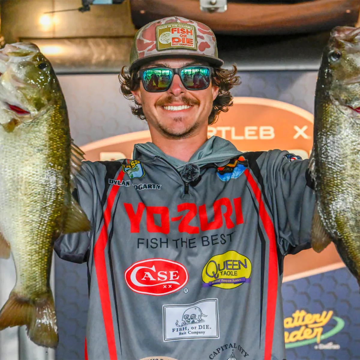 DYLAN FOGARTY: Wearing a gray sponsor jersey, sunglasses, and a cap holds two large bass fish outstretched on both sides. The background shows a branded event wall with 'Fish or Die' and other sponsor logos.