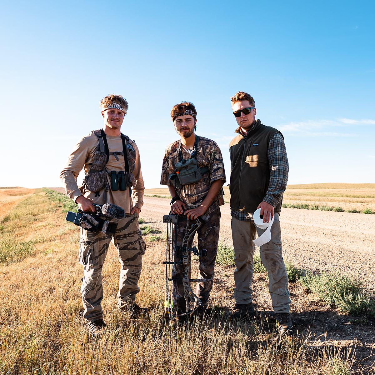 GONE ALL SEASON: Three team members in outdoor hunting apparel and gear stand in a grassy field under a clear blue sky. The center man holds a compound bow, while the others carry or wear hunting accessories including vests, sunglasses, and a camera.