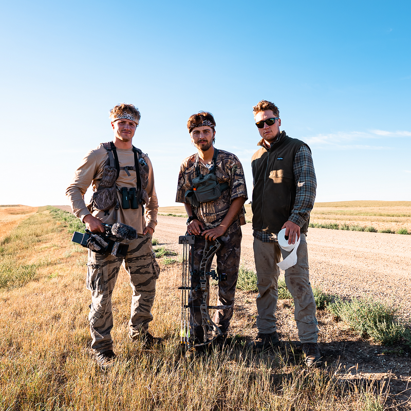 GONE ALL SEASON: Three team members in outdoor hunting apparel and gear stand in a grassy field under a clear blue sky. The center man holds a compound bow, while the others carry or wear hunting accessories including vests, sunglasses, and a camera.