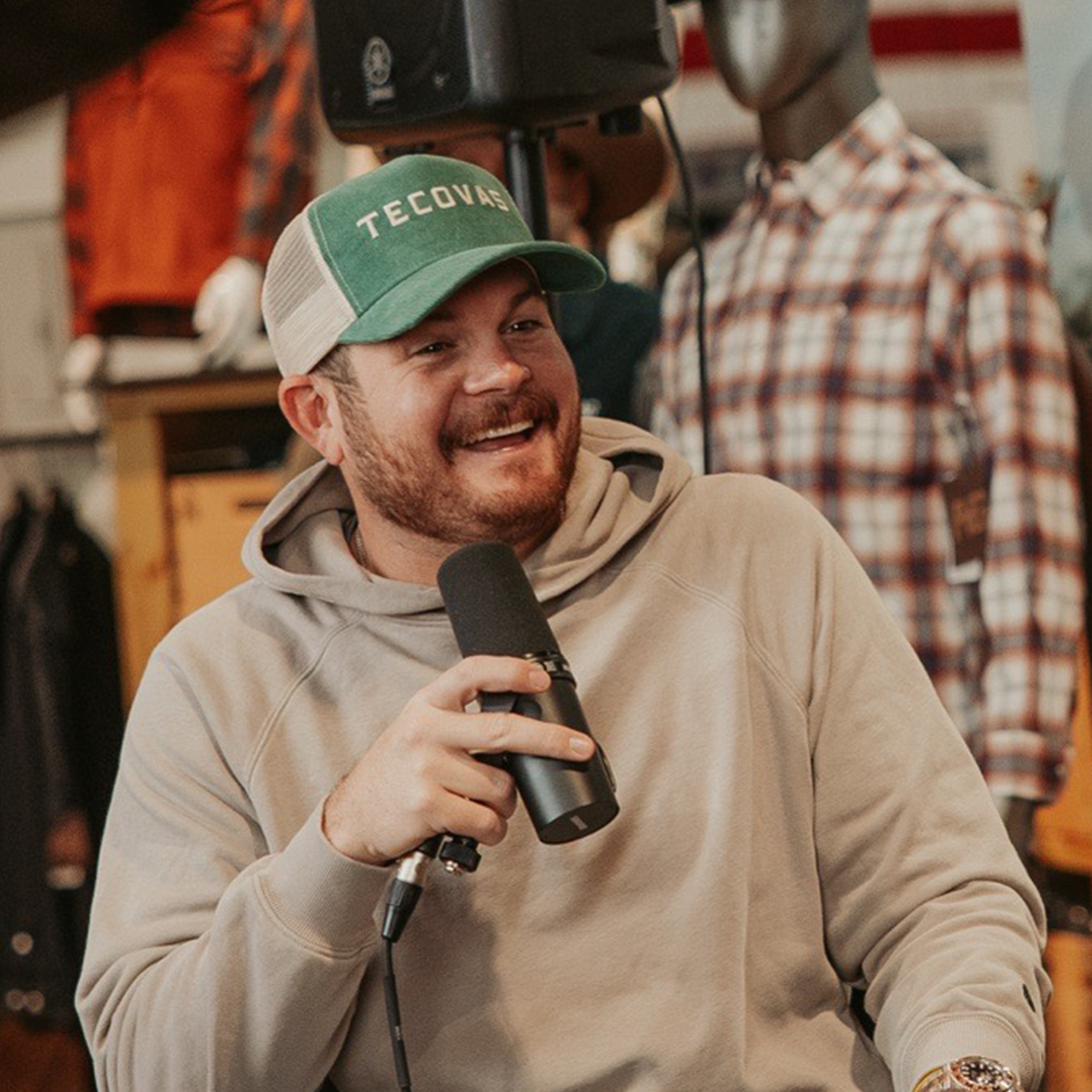 LIKE A FARMER, PAT SPINOSA: In a beige hoodie and green trucker hat, holding a black microphone in a retail store setting with mannequins in the background.