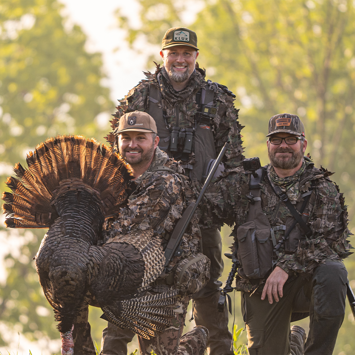 UNTAMED: Three team members in camouflage hunting apparel pose outdoors with a large wild turkey in a wooded setting. One man wears a full ghillie suit, another kneels holding the turkey, and the third stands behind holding a rifle.
