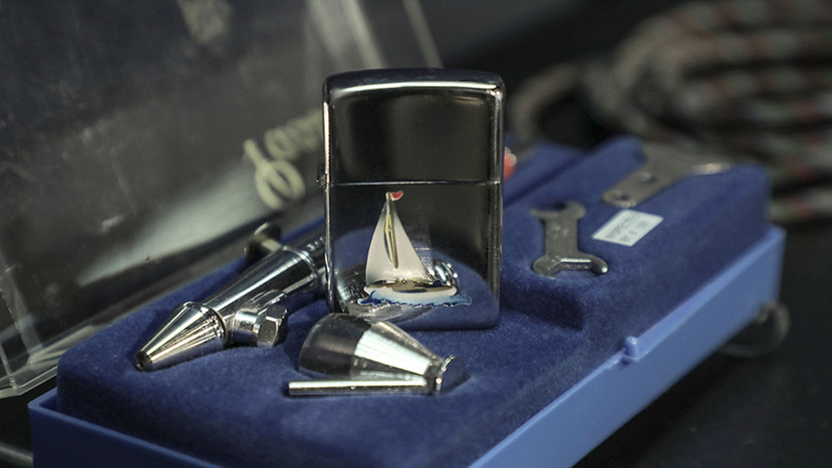 A polished silver metal lighter with a miniature sailboat design on the front, displayed in a blue velvet-lined case alongside matching metal tools or accessories.