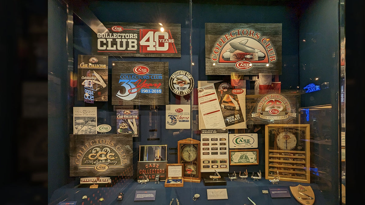 A glass display case showcasing a collection of vintage and modern Collectors Clu signs, plaques, trophies, and memorabilia against a dark blue background.