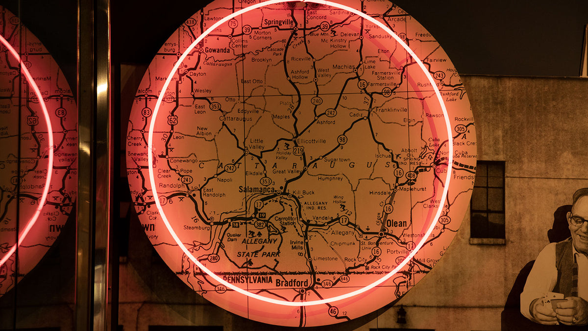 A circular vintage-style map neon sign glowing with a red-pink light, mounted on a dark wall next to a sepia-toned portrait.