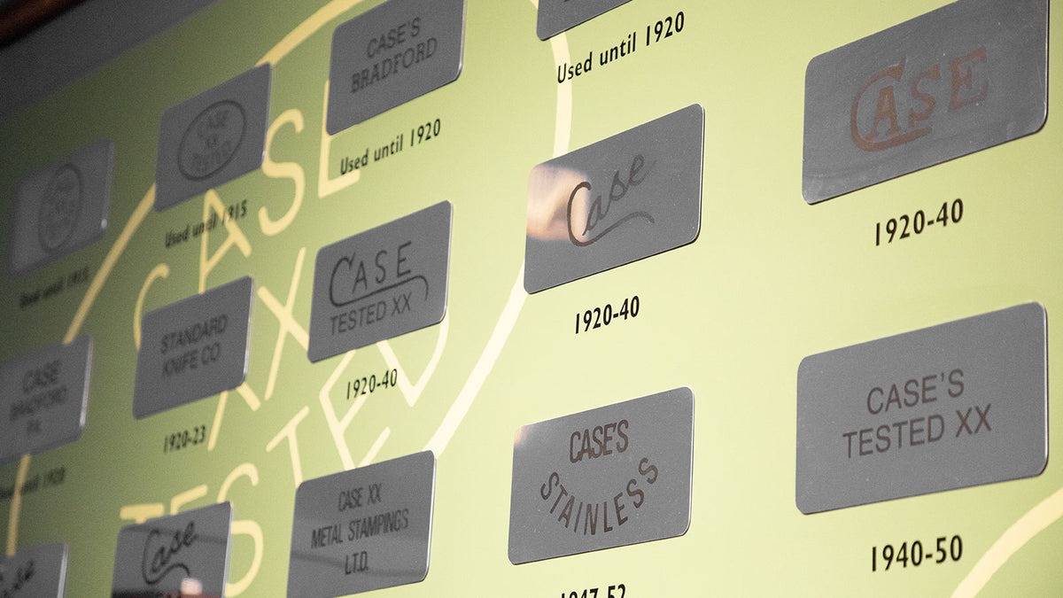 A close-up of a vintage display wall featuring multiple gray rectangular plaques with engraved text such as 'CASE', 'CASE'S BRADFORD', 'CASE'S STAINLESS', and dates like '1920-40' and '1940-50' on a pale green background.