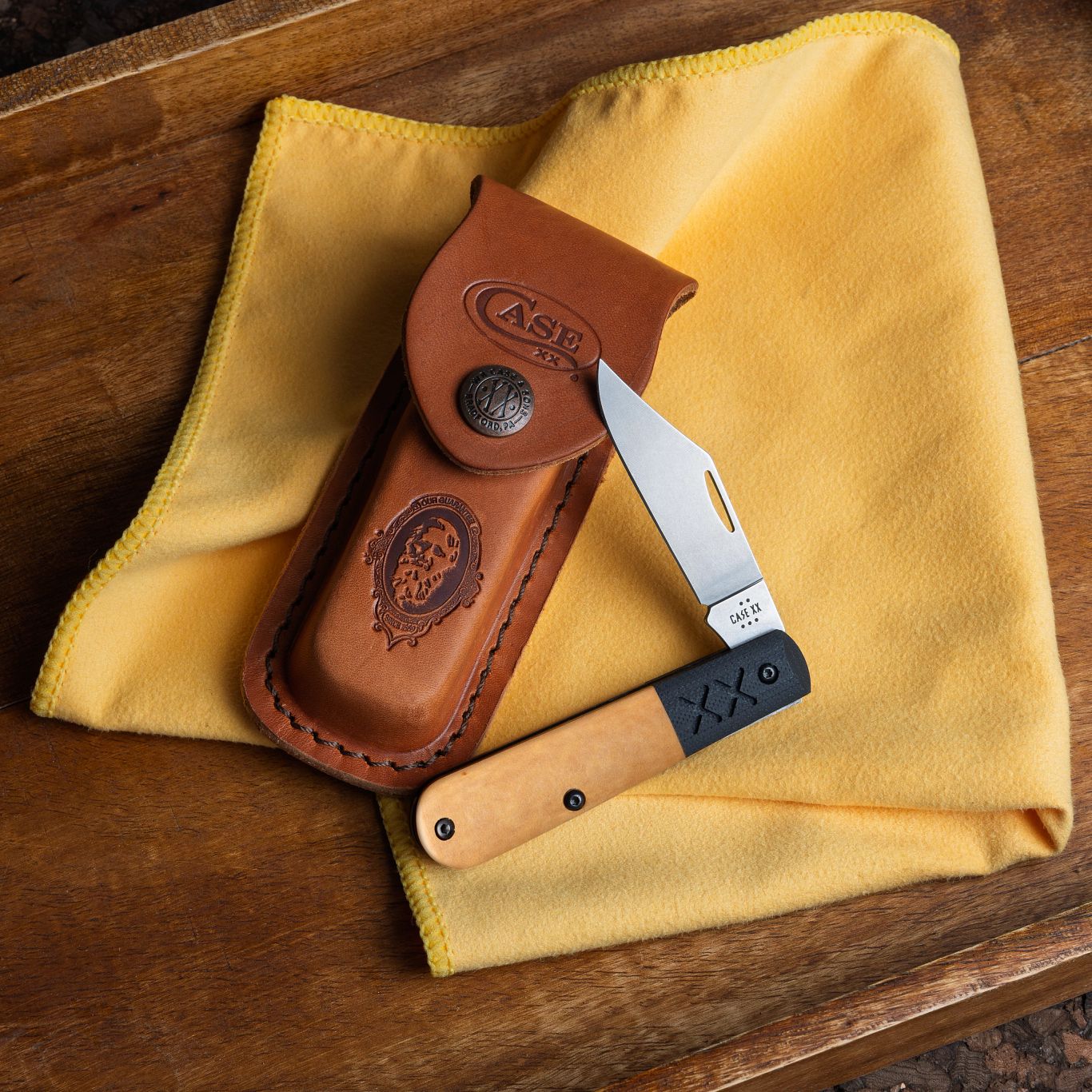 Lifestyle image of Crossroads Smooth Tan Richlite® w/Black G-10 XR Barlow open and resting on a sheath with a cloth and wooden surface underneath.