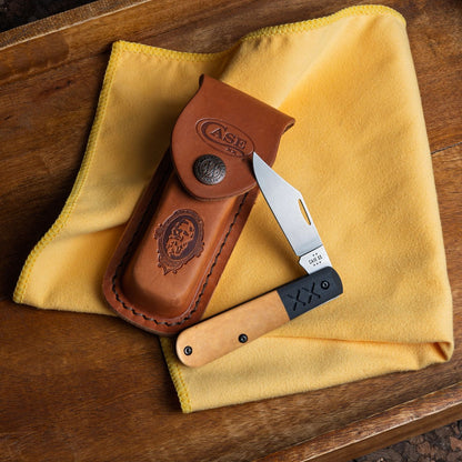 Lifestyle image of Crossroads Smooth Tan Richlite® w/Black G-10 XR Barlow open and resting on a sheath with a cloth and wooden surface underneath.