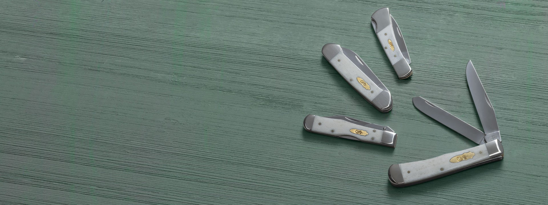 Four Case knives with silver blades and handles, featuring a gold-toned logo emblem, arranged on a textured green wooden surface.