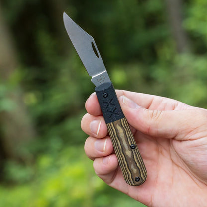 Lifestyle image of Crossroads Smooth Black & Tan Richlite® w/Black G-10 XR Barlow open and held in hand with a blurred forest scene in the background.